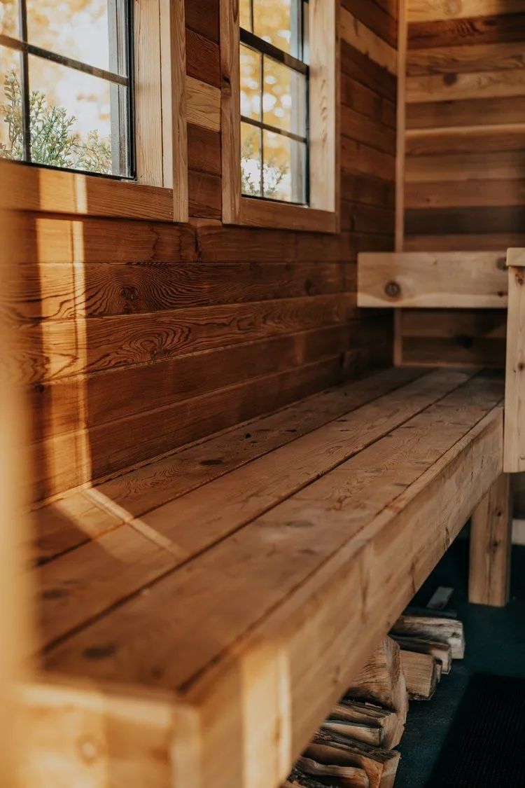 Wooden bench in a cabin interior, sunlight through windows. Wood-paneled walls and a stack of firewood visible.