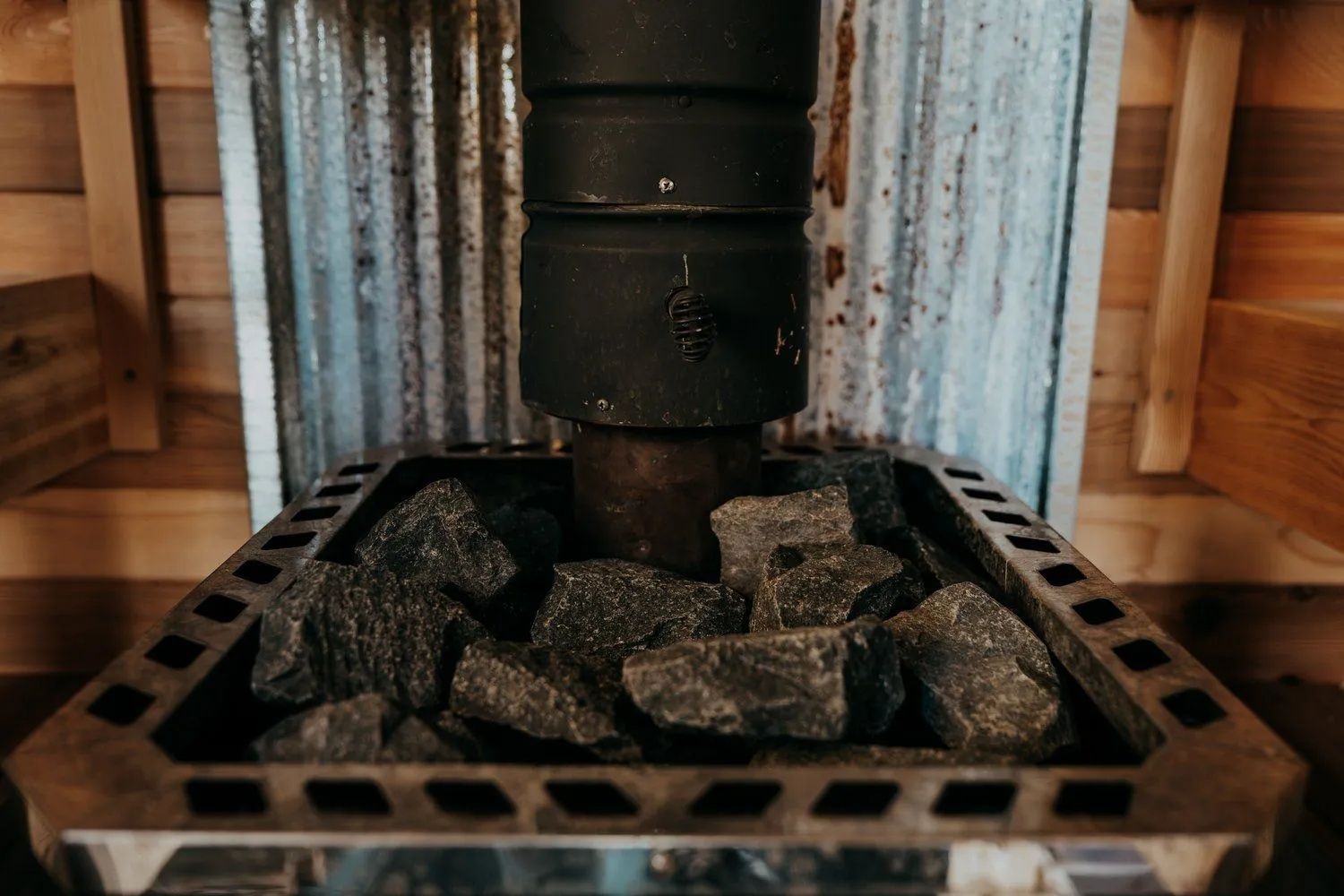 Sauna heater with dark rocks and a black chimney. Wooden walls and corrugated metal back.