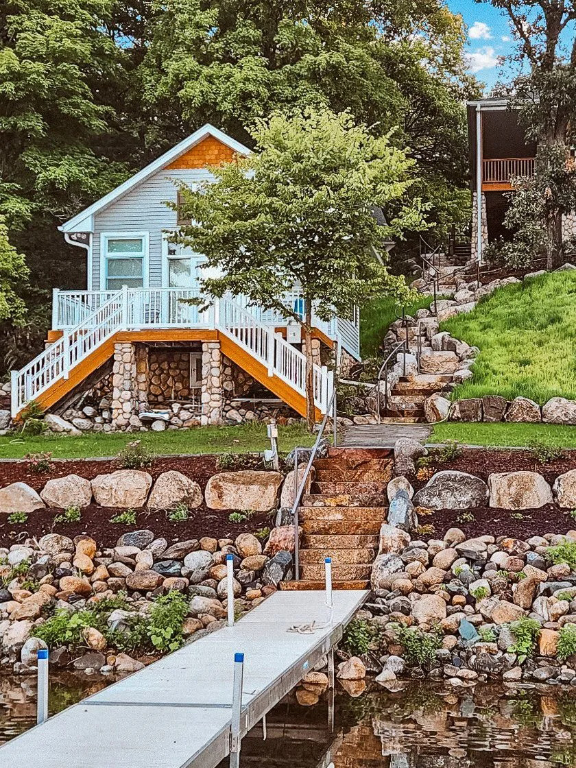 Cottage with dock on lake. White and orange trim, rock retaining walls, trees, blue chairs.