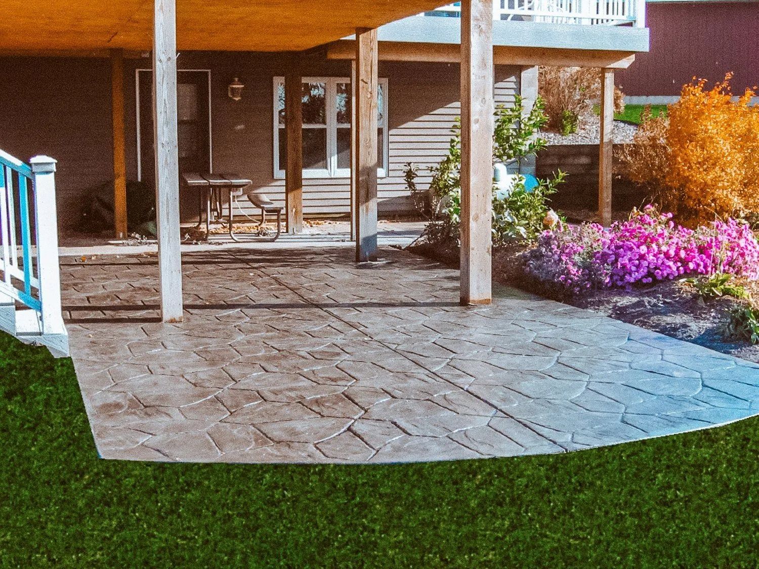 Stamped concrete patio with a deck overhead, surrounded by green grass and flowers.
