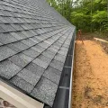Gabled roof with gray shingles and a black gutter beside a brick wall and wooded yard.