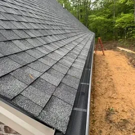 Gabled roof with gray shingles and a black gutter beside a brick wall and wooded yard.
