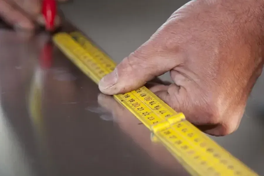 Hand measuring a metal surface with a yellow tape measure and marking it with a red pencil