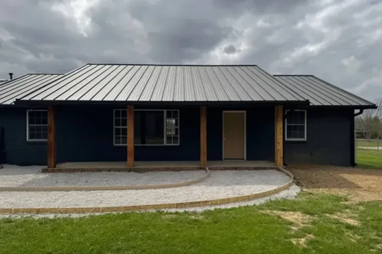 Single-story dark house with metal roof, front porch, and cloudy sky over a grassy yard