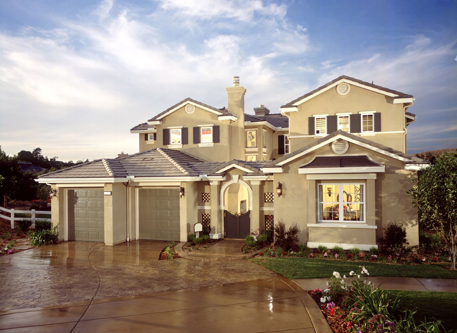 Two-story beige house with attached garage, driveway, and landscaping under a sunny sky.