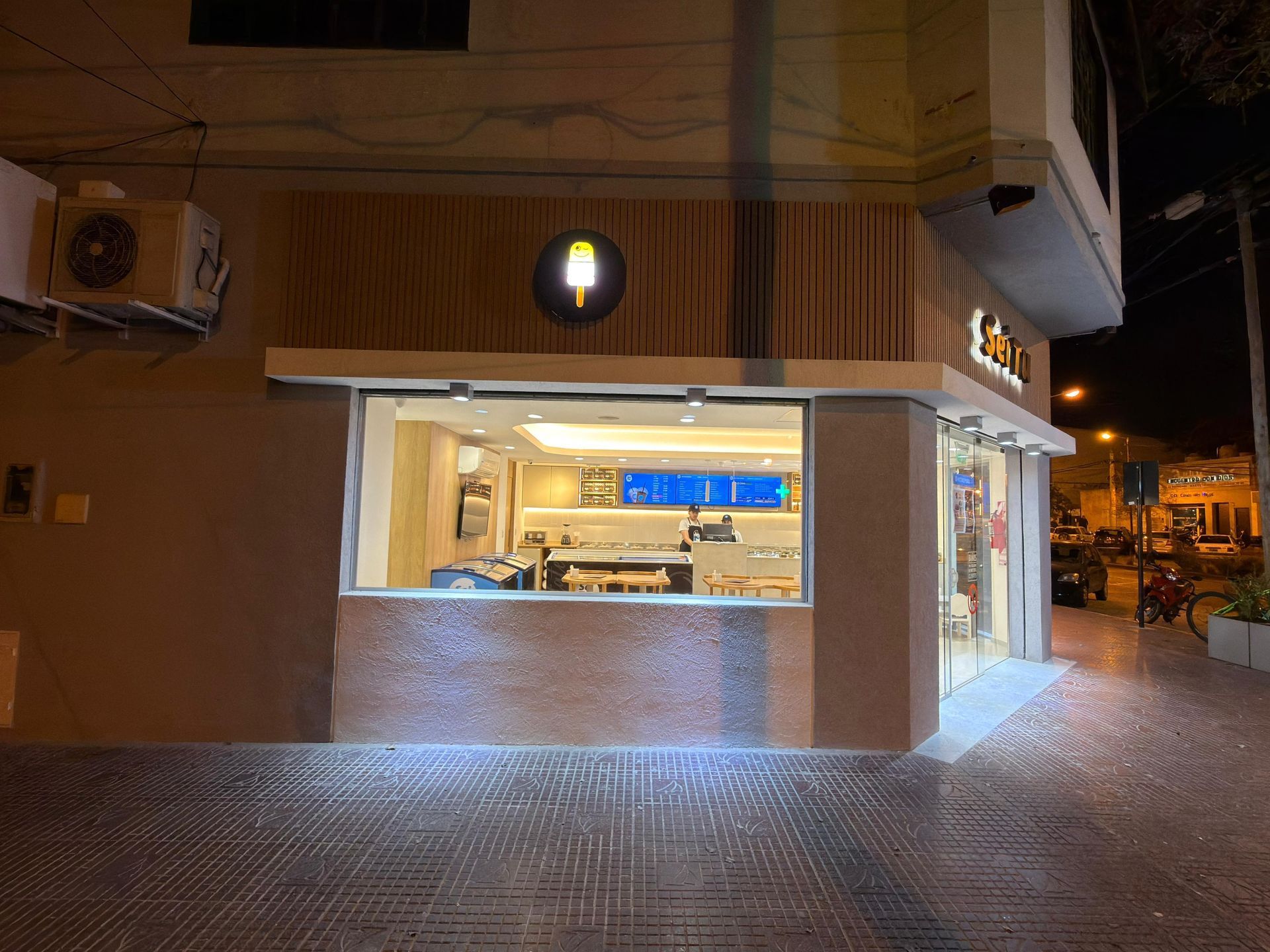 Vista nocturna de una cafetería iluminada en una esquina, con un gran ventanal y una terraza en la acera.