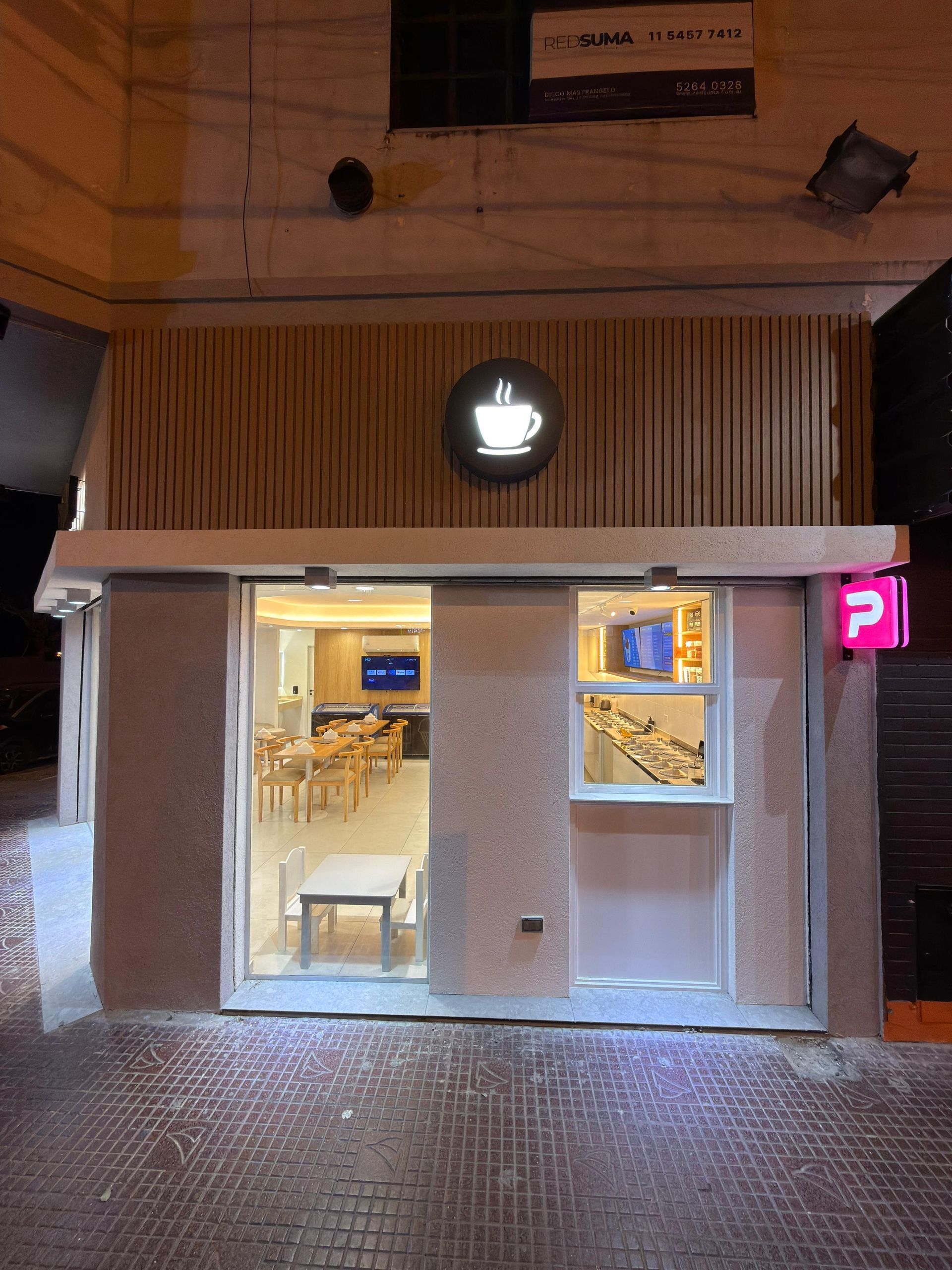 Pequeña cafetería con fachada iluminada por la noche, interior blanco y letrero de neón rosa.