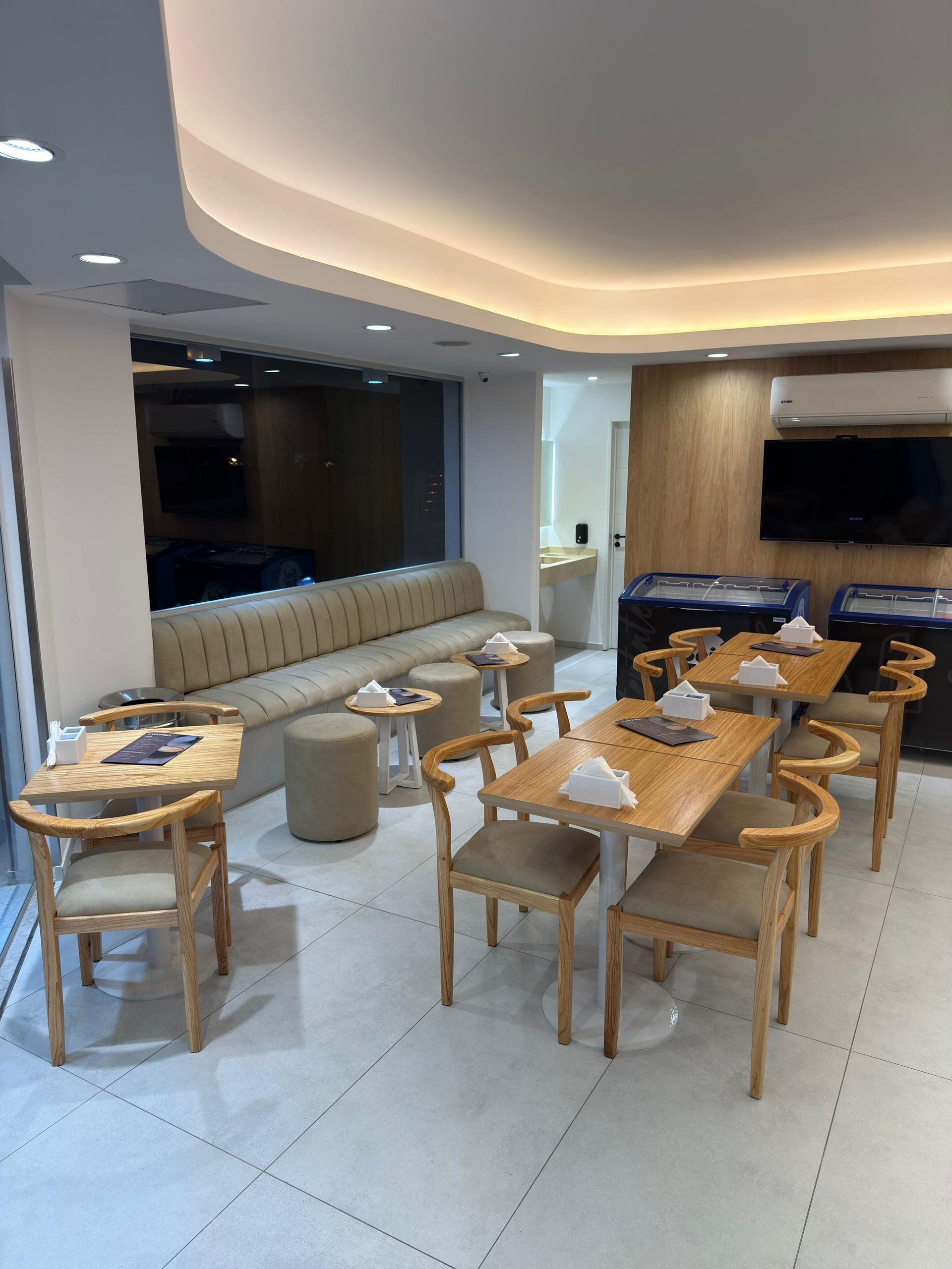 Cafetería moderna con mesas de madera, taburetes, asientos tipo banqueta capitoné y una cálida iluminación en el techo.