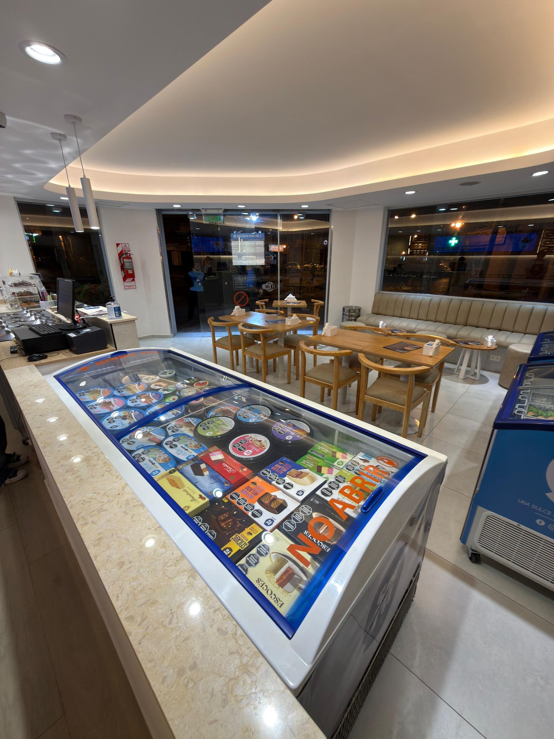 Mostrador de sushi en un restaurante muy iluminado con mesas y sillas de madera al fondo.