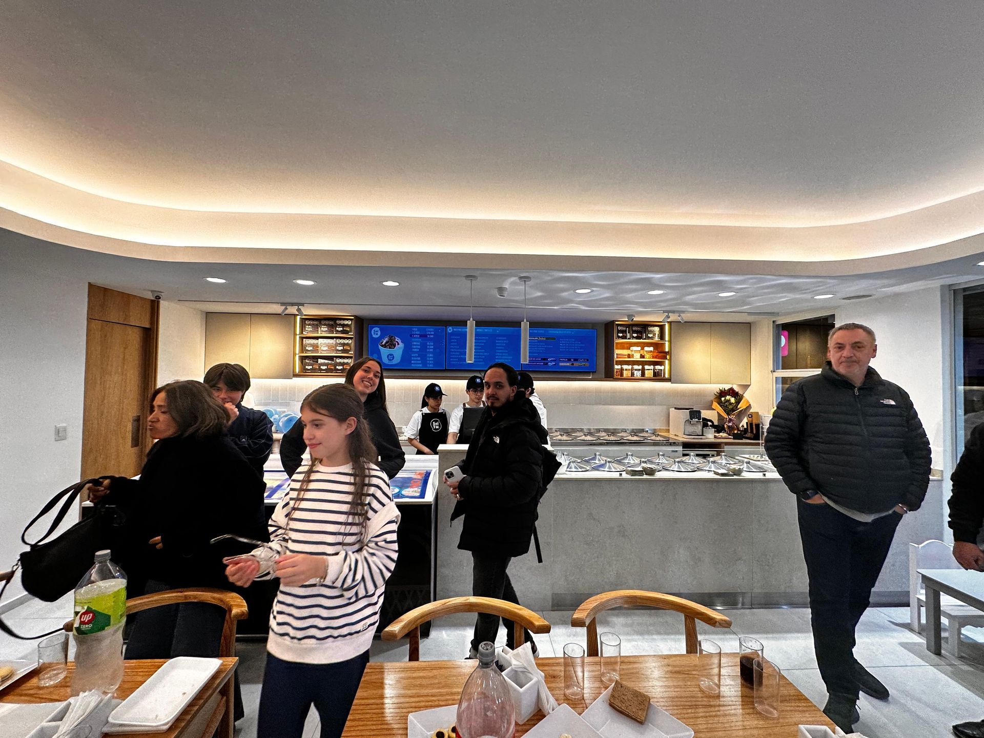Gente en una cafetería o panadería luminosa, con un mostrador, pizarras con el menú y mesas en primer plano.