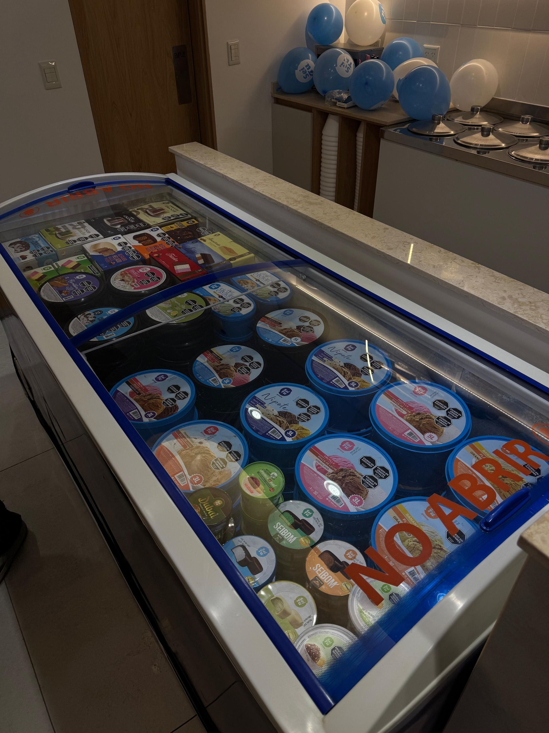 Congelador expositor de helados con coloridos recipientes bajo una tapa de cristal en una tienda.