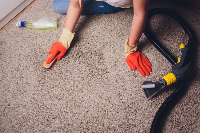 Person in orange gloves vacuuming carpet beside a bottle on the floor