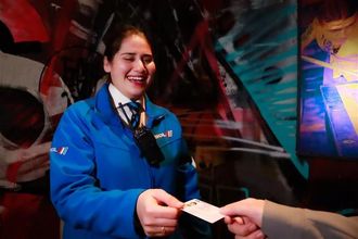 A smiling person in a blue uniform hands a small white card to someone against a background of colorful wall art.