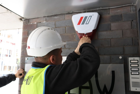 A worker in a hard hat and high-visibility vest installs or maintains a white alarm box on a brick building exterior.