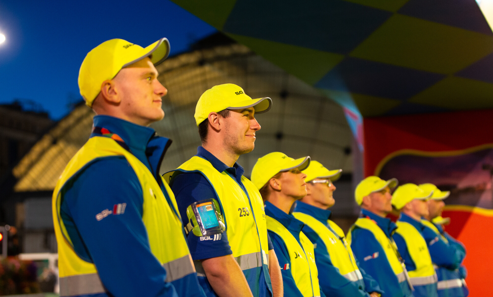 A line of people in yellow caps and high-visibility yellow and blue vests stand outdoors, facing the same direction.