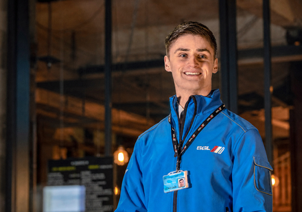 A person in a blue jacket with a lanyard stands in front of a brightly lit 
