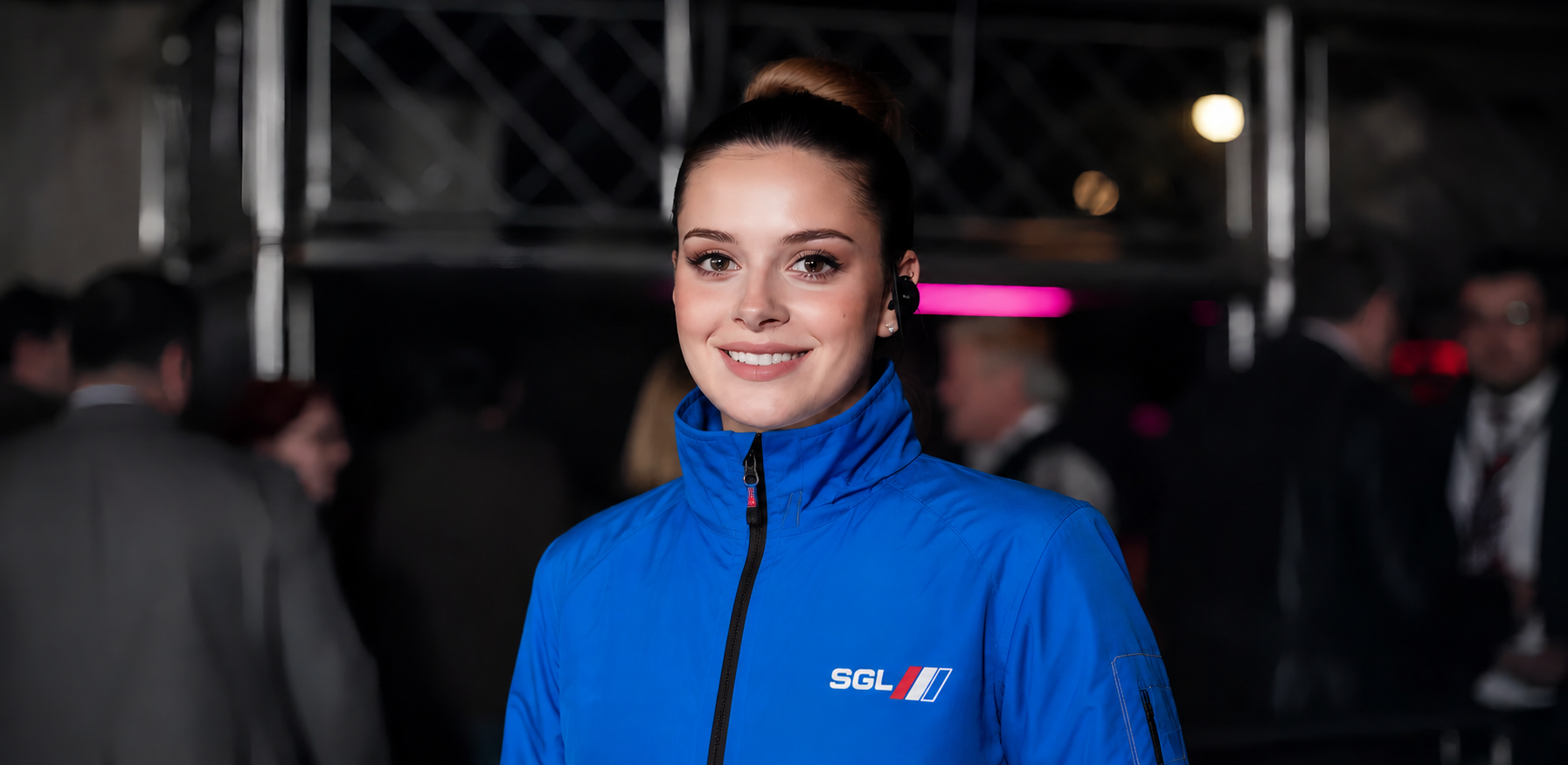 Five people in matching black jackets with logos stand side-by-side, smiling, in front of a blurry, light-streaked city.