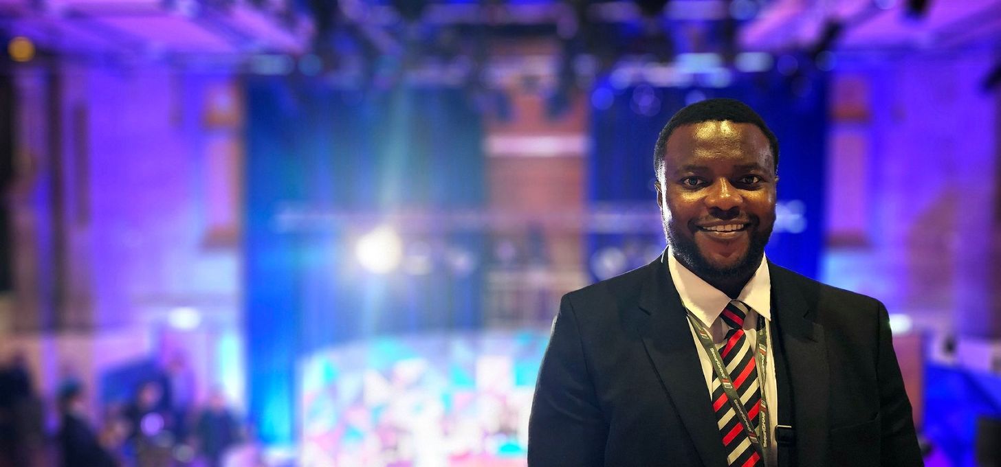 A person in a suit and striped tie smiles in front of a blurry, blue-lit event space.