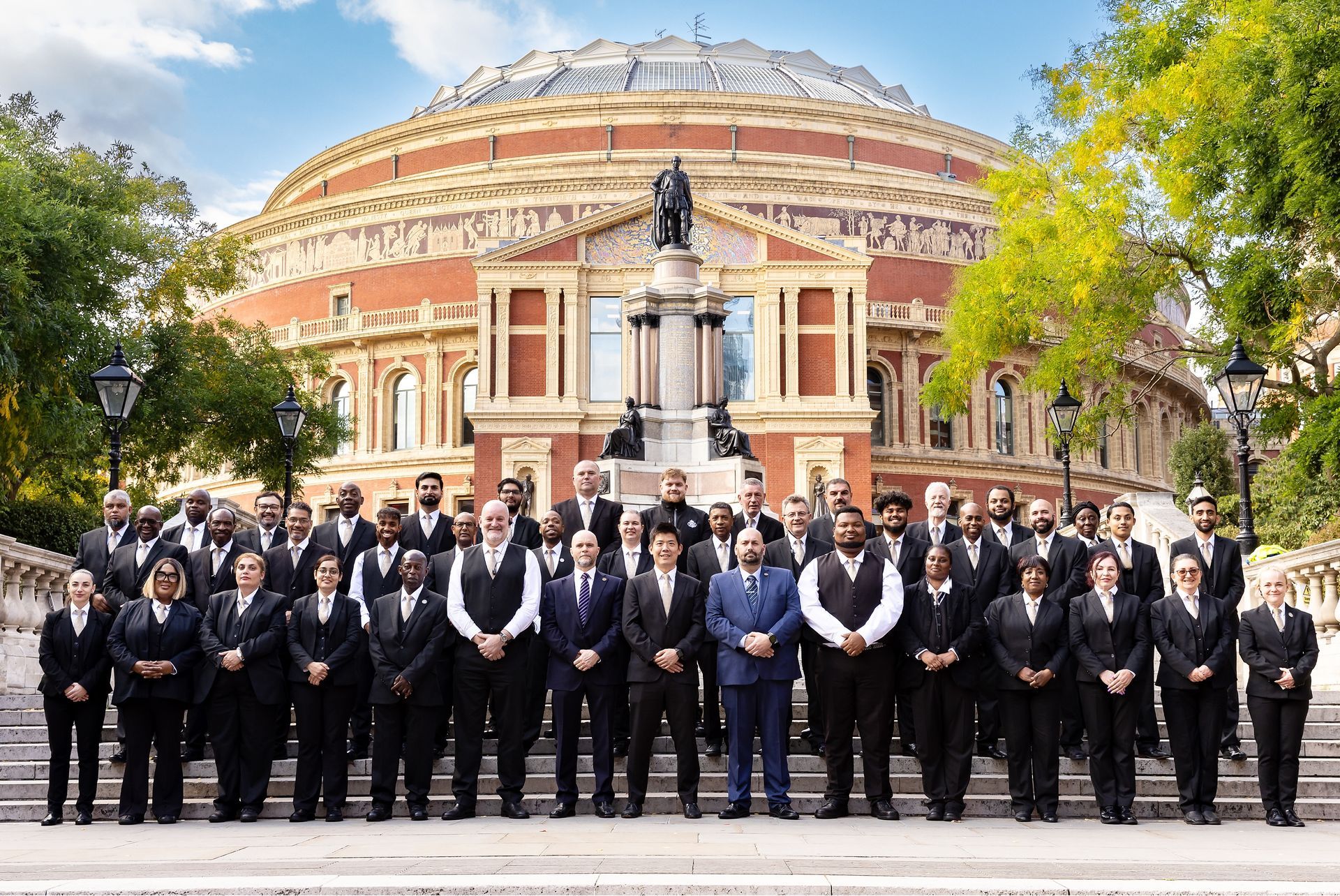 A large group of people in suits stands on the steps in front of the circular Royal Albert Hall on a sunny day.