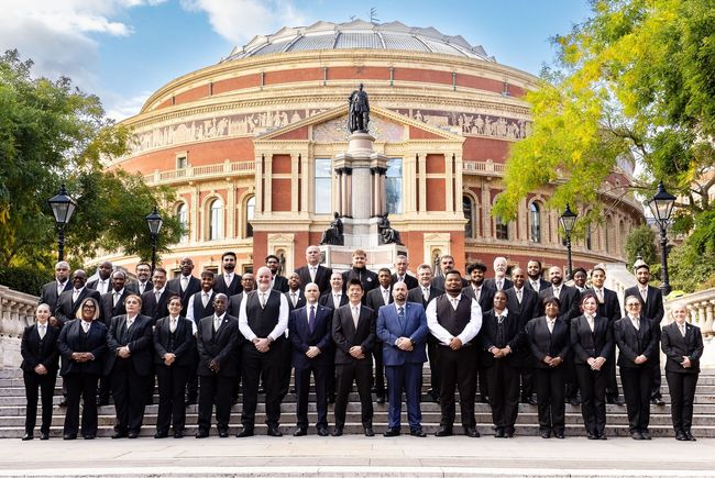 A large group of people in suits stands on the steps in front of the circular Royal Albert Hall on a sunny day.