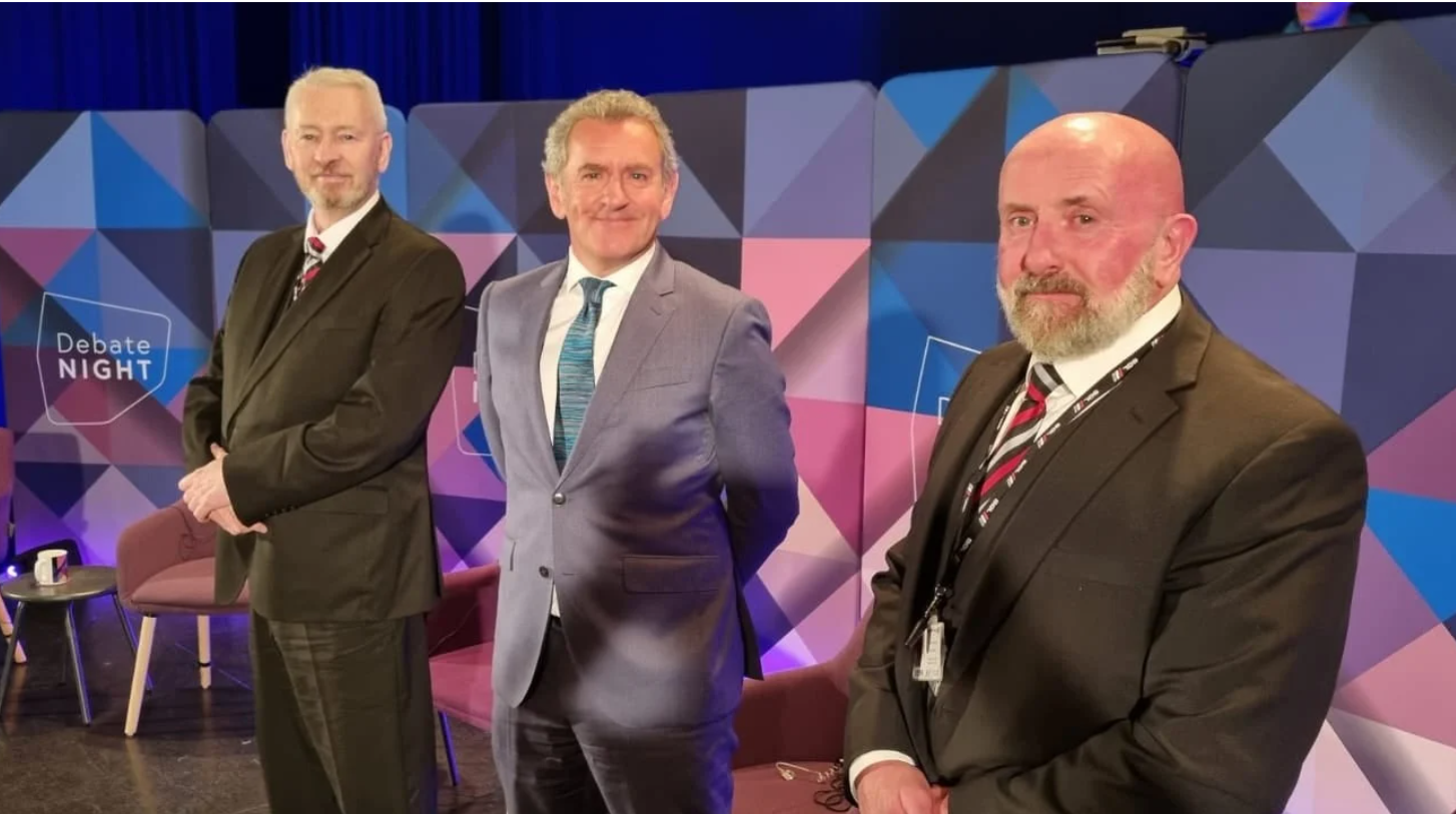 Three men in suits stand on a stage with a geometric blue, purple, and pink backdrop, positioned for a television debate.