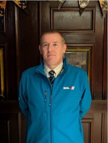 A man wearing a teal branded zip-up jacket and a collared shirt with a tie stands before a dark, paneled door.