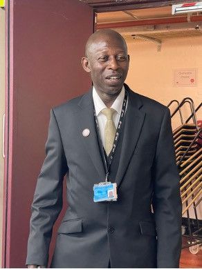 A person in a suit and tie with a security lanyard standing in a doorway with stacked chairs in the background.