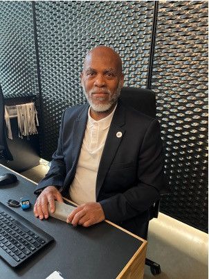 A person wearing a suit jacket and collared shirt sits at a desk with a computer keyboard, facing the camera.