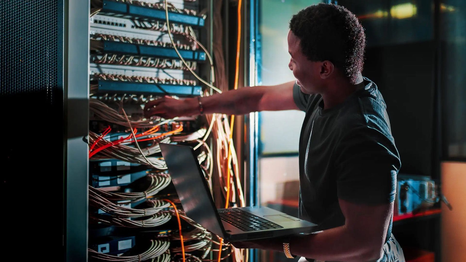A man is working on a laptop in a server room.
