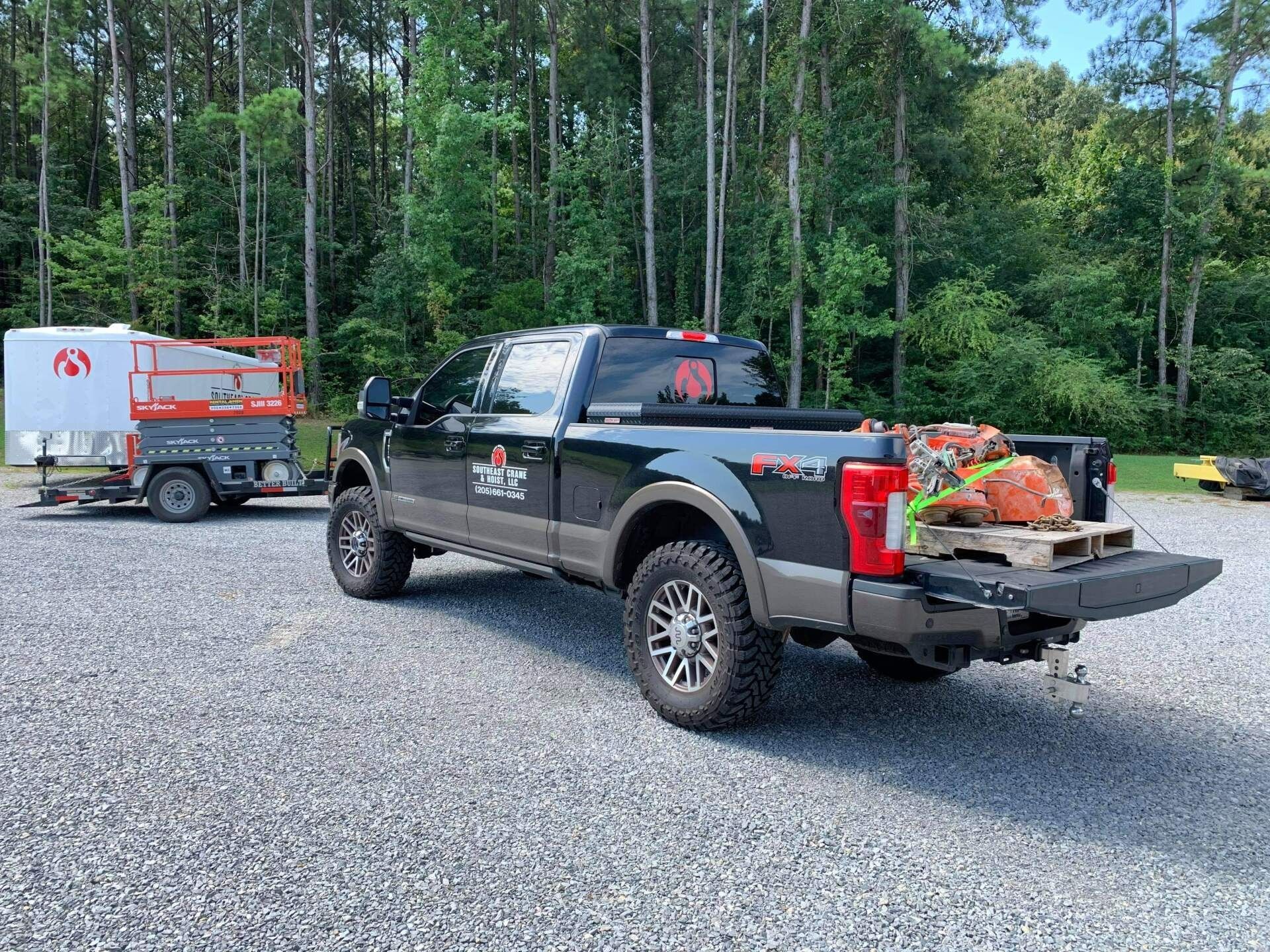 Dark pickup truck towing a trailer with a lift on gravel, tools in the truck bed, trees in the background.