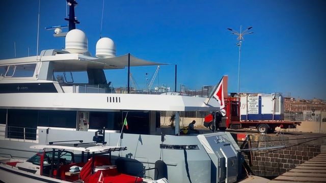 Un grande yacht bianco ormeggiato in porto accanto a camion rossi e gru, sotto un cielo azzurro e terso
