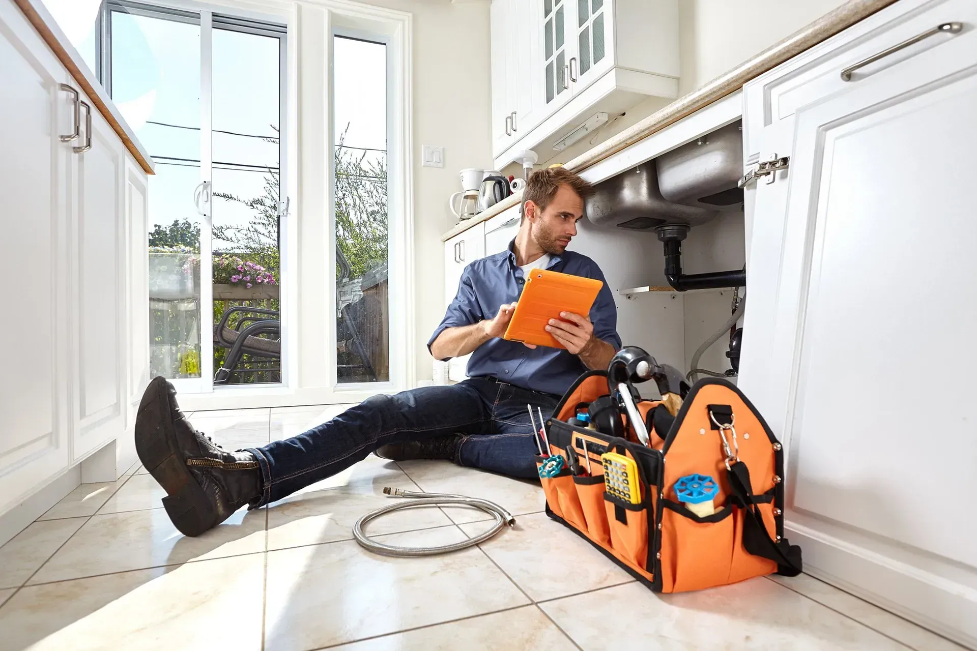 A man is sitting on the floor in a kitchen looking at a tablet.
