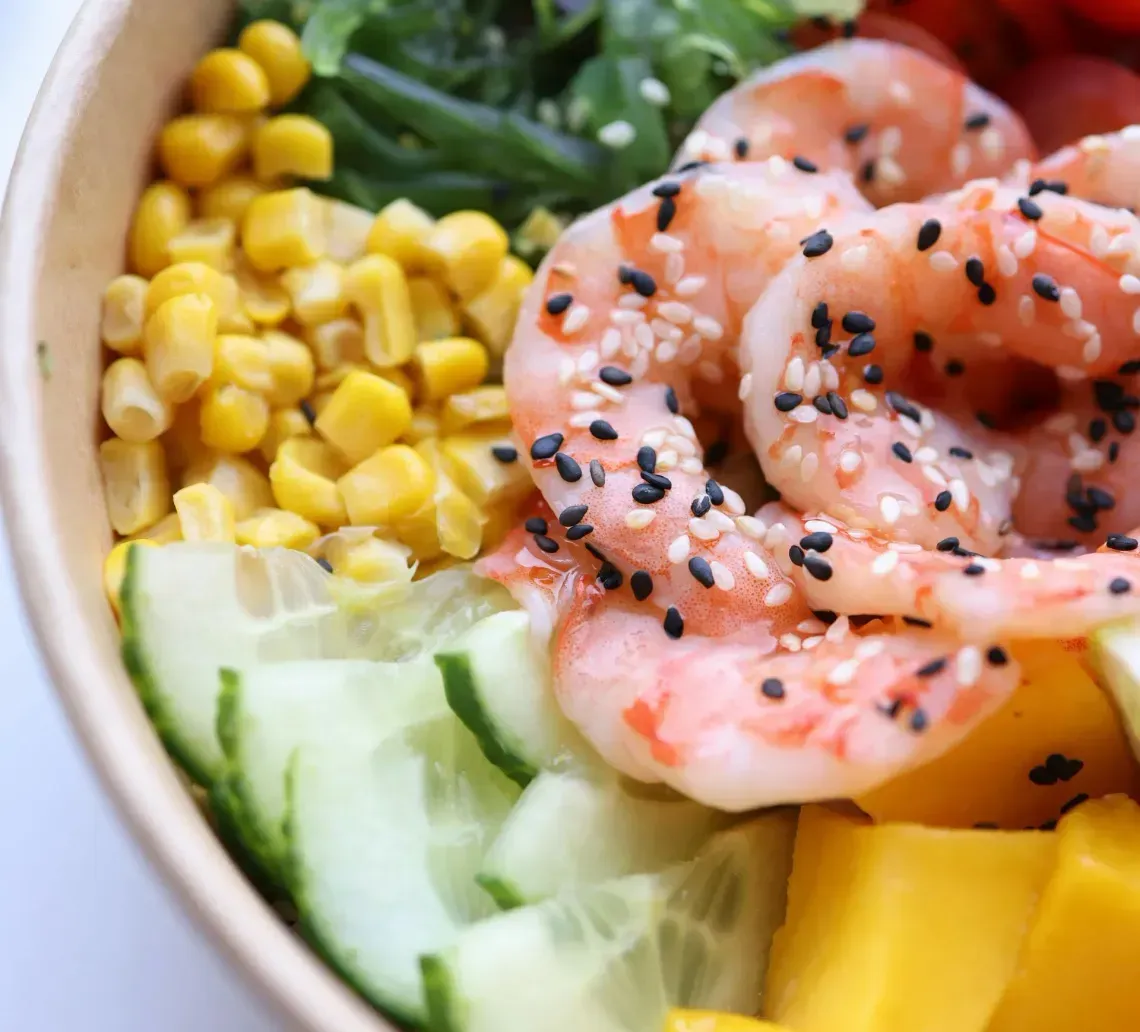 A colorful poke bowl with shrimp, corn, cucumber, mango, greens, and cherry tomatoes topped with sesame seeds