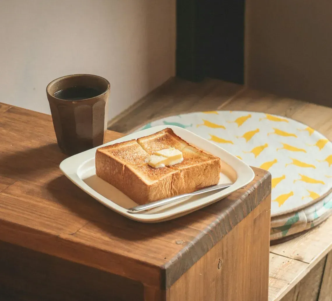 Toast on a plate beside a cup on a wooden table, with a patterned cushion in the background.