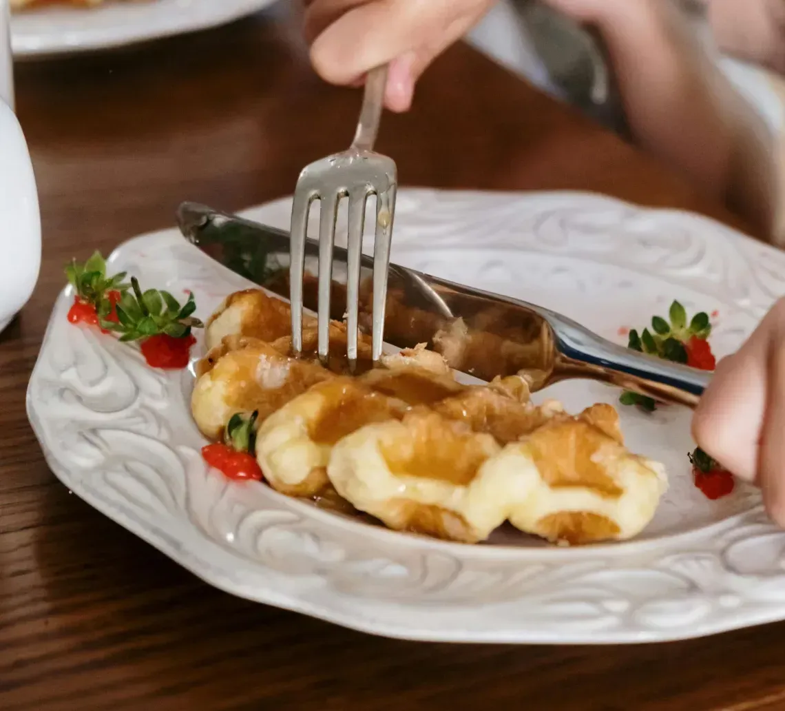 Hands serving food on a white plate at a wooden table with tomatoes and greens