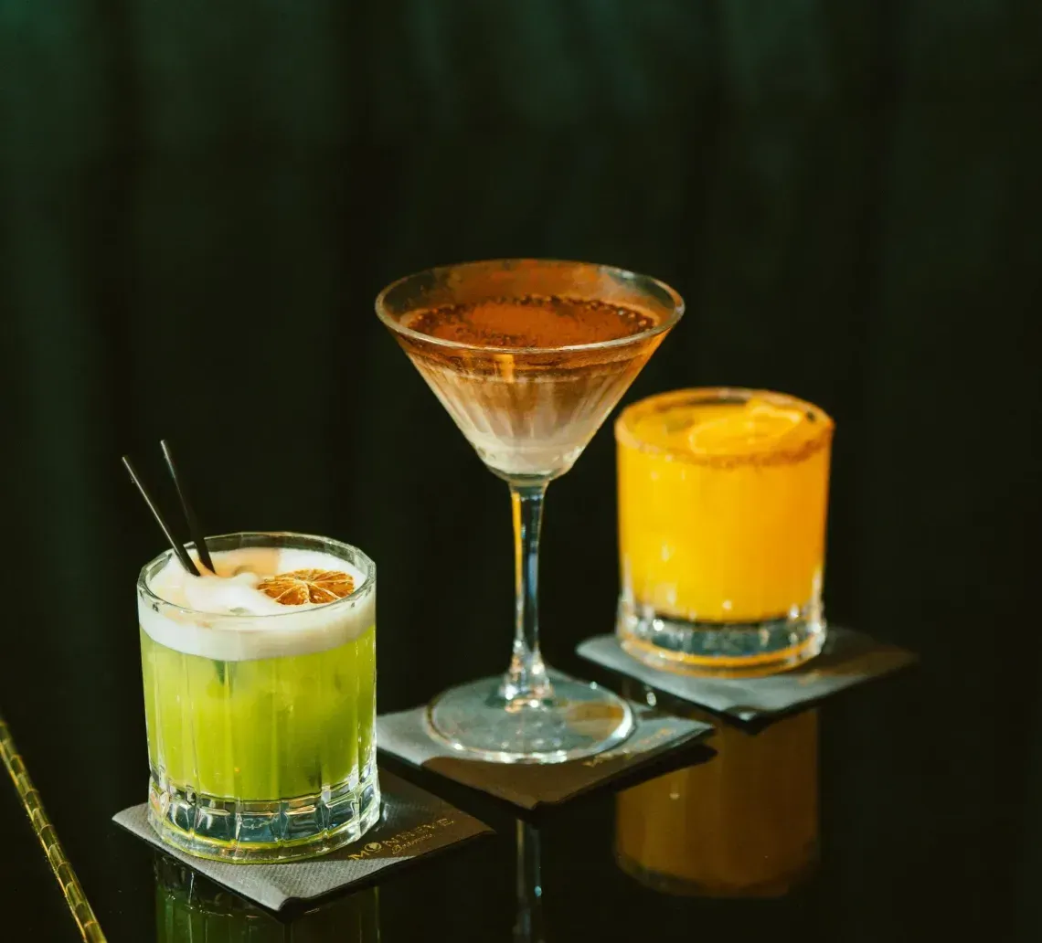 Three colorful cocktails on a glossy black table, with a dark green backdrop.