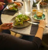 People sharing a meal at a round table with salads, drinks, and bread in a warm café setting