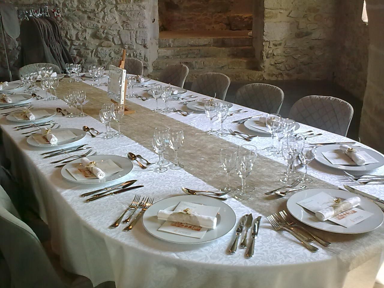 Une longue table de banquet dressée avec des assiettes blanches, des verres et des couverts en argent dans une salle à manger rustique.