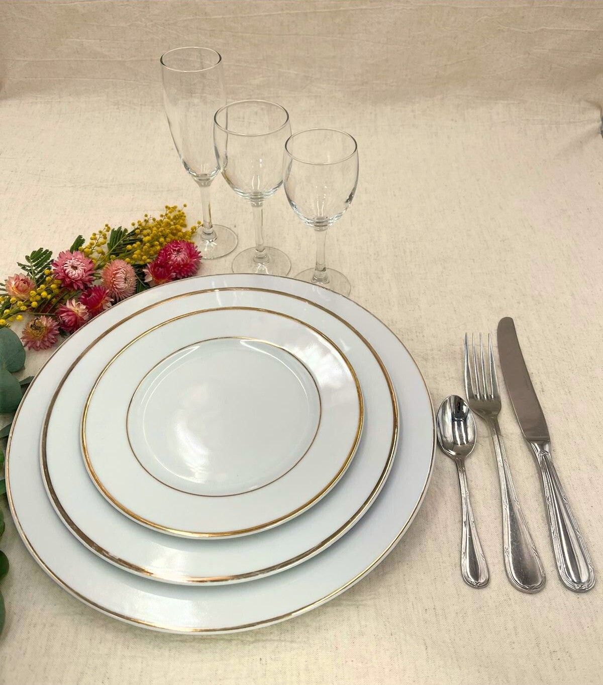 Élégante table dressée avec assiettes bordées d'or blanc, couverts, verres et petites fleurs sur une nappe beige