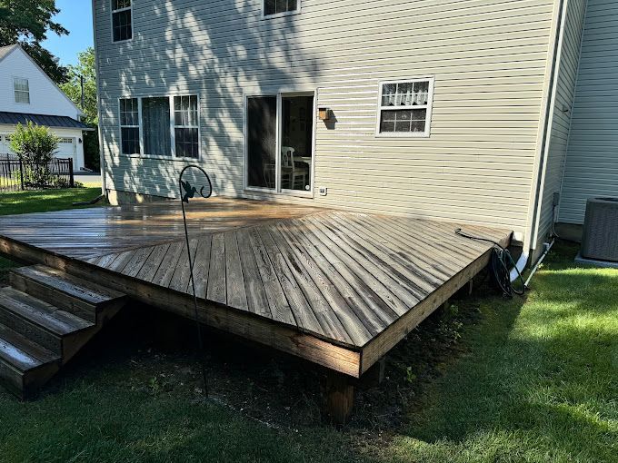 Wooden deck attached to a two-story beige house. The deck is wet, with steps leading down to the lawn