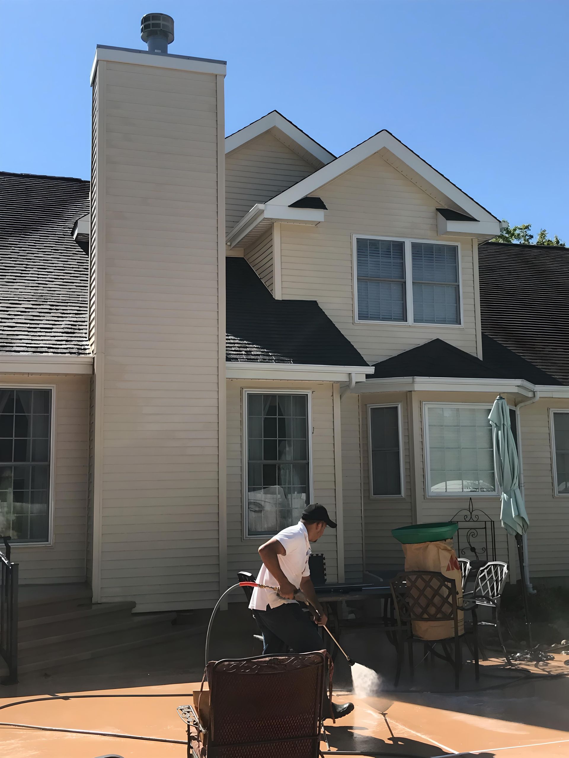 Person pressure washing a backyard patio next to a two-story beige house with a chimney on a sunny day