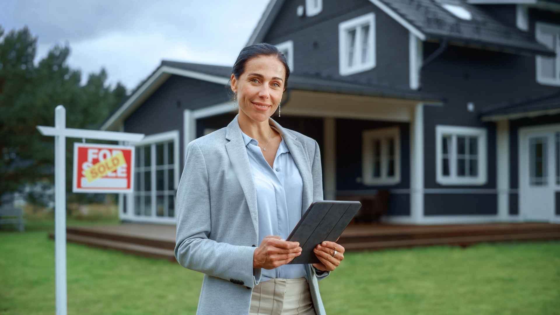 A woman is standing in front of a house holding a tablet.