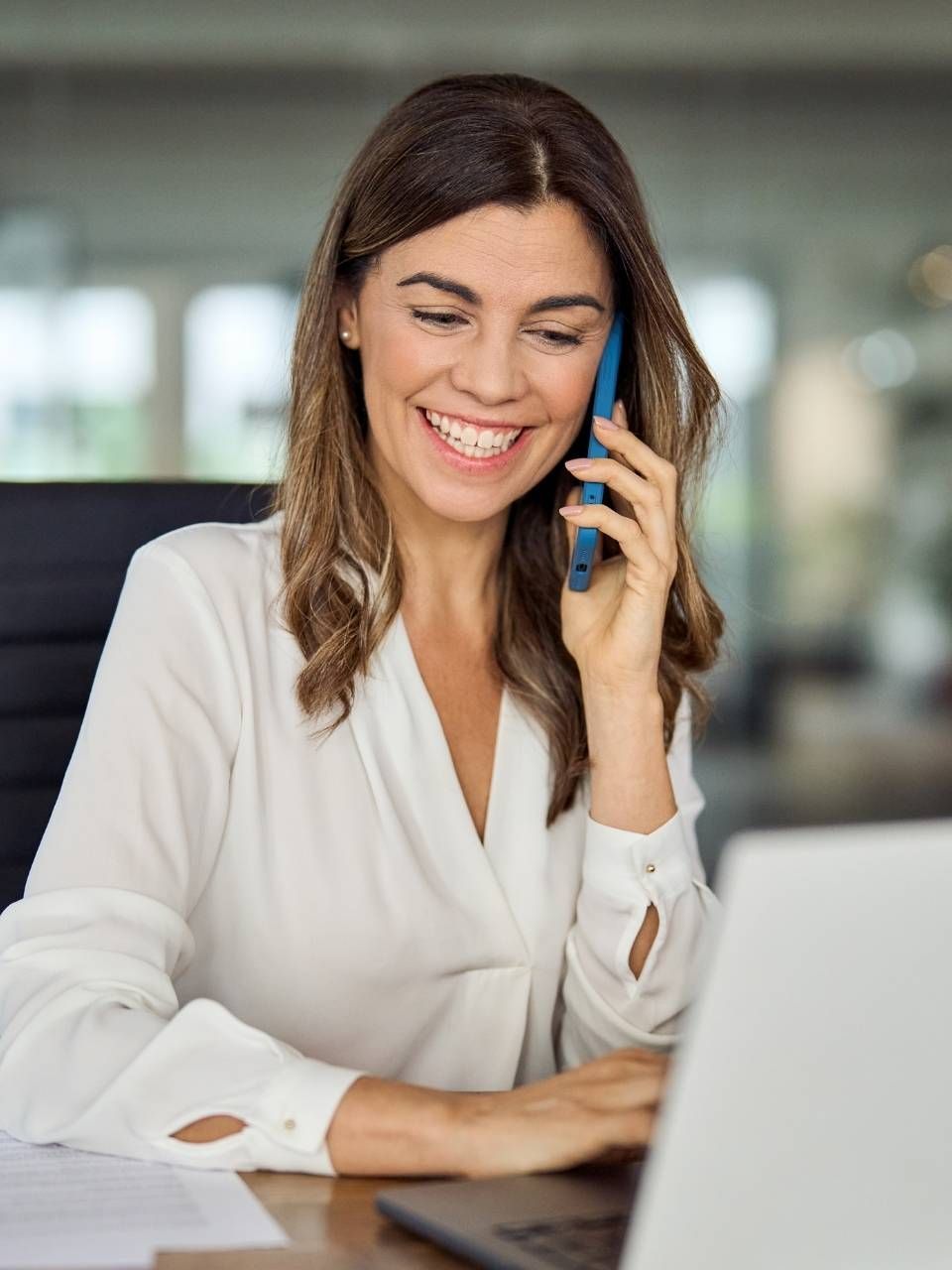 A woman is talking on a cell phone while using a laptop computer.