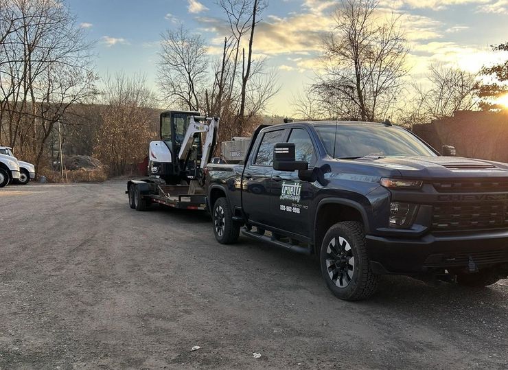 Black pickup truck towing a small lift on a gravel lot at sunset with bare trees in the background