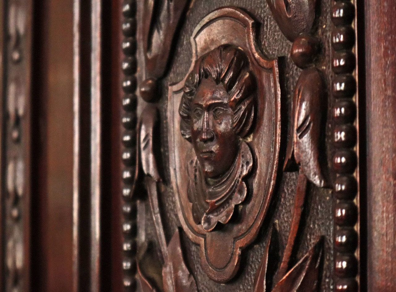 Panel de madera tallada con el retrato del rostro de una persona enmarcado por hojas y adornos decorativos.