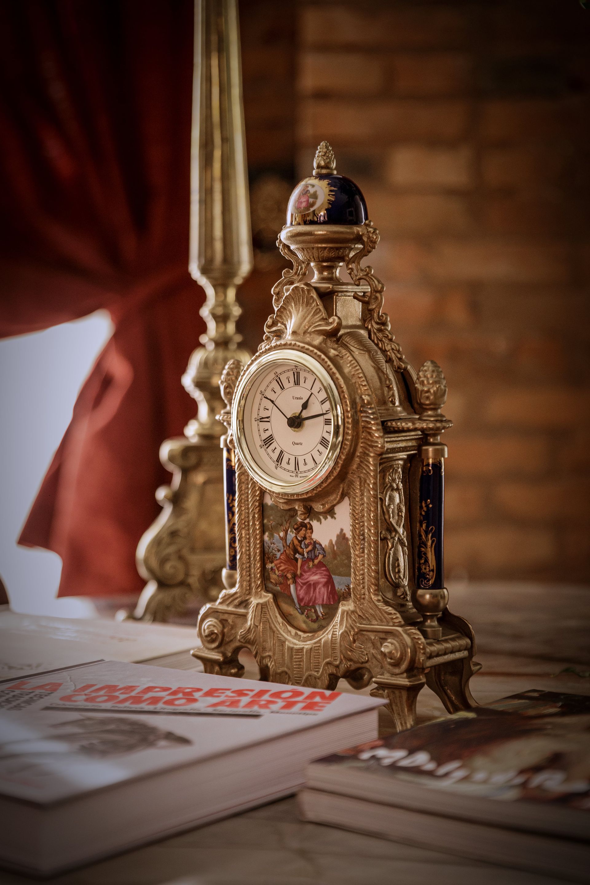 Reloj dorado adornado sobre una mesa con libros y un candelabro contra una cortina roja y una pared de ladrillo.