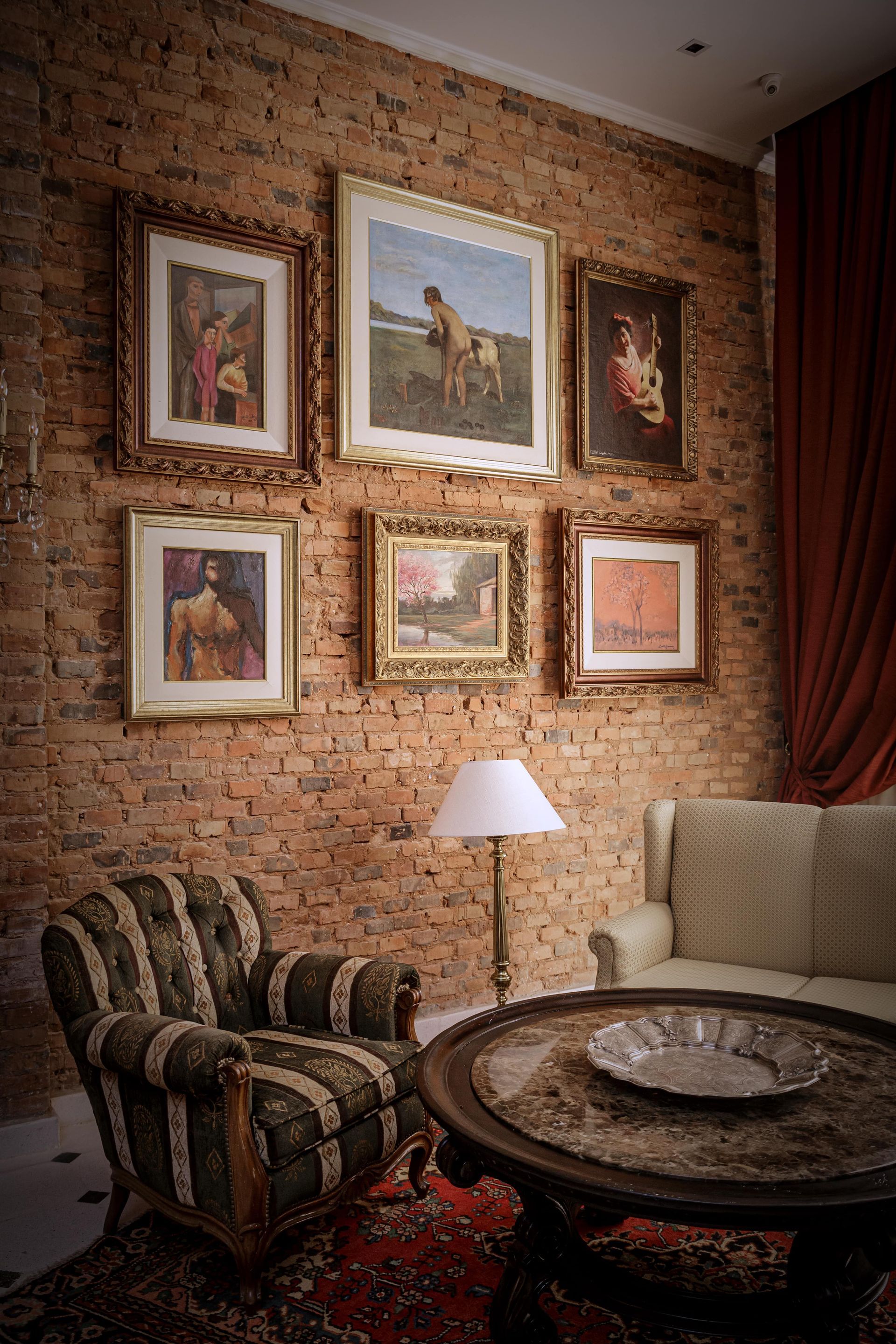Pared de galería con cuadros enmarcados sobre pared de ladrillo, encima de un sillón a rayas, un sofá y una mesa redonda.