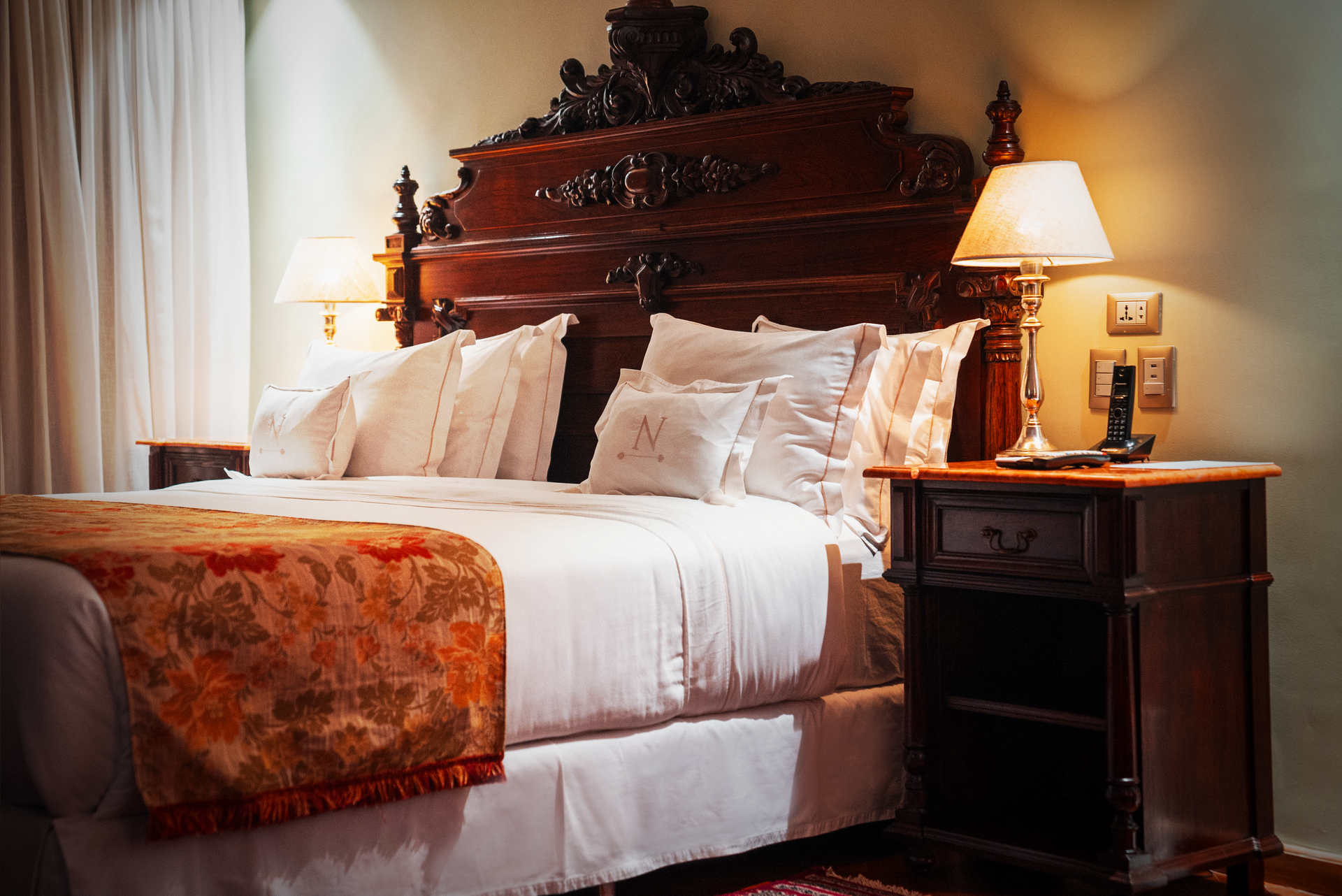 Una acogedora habitación de hotel con un cabecero grande y ornamentado de madera tallada, ropa de cama blanca y una mesita de noche con lámpara.