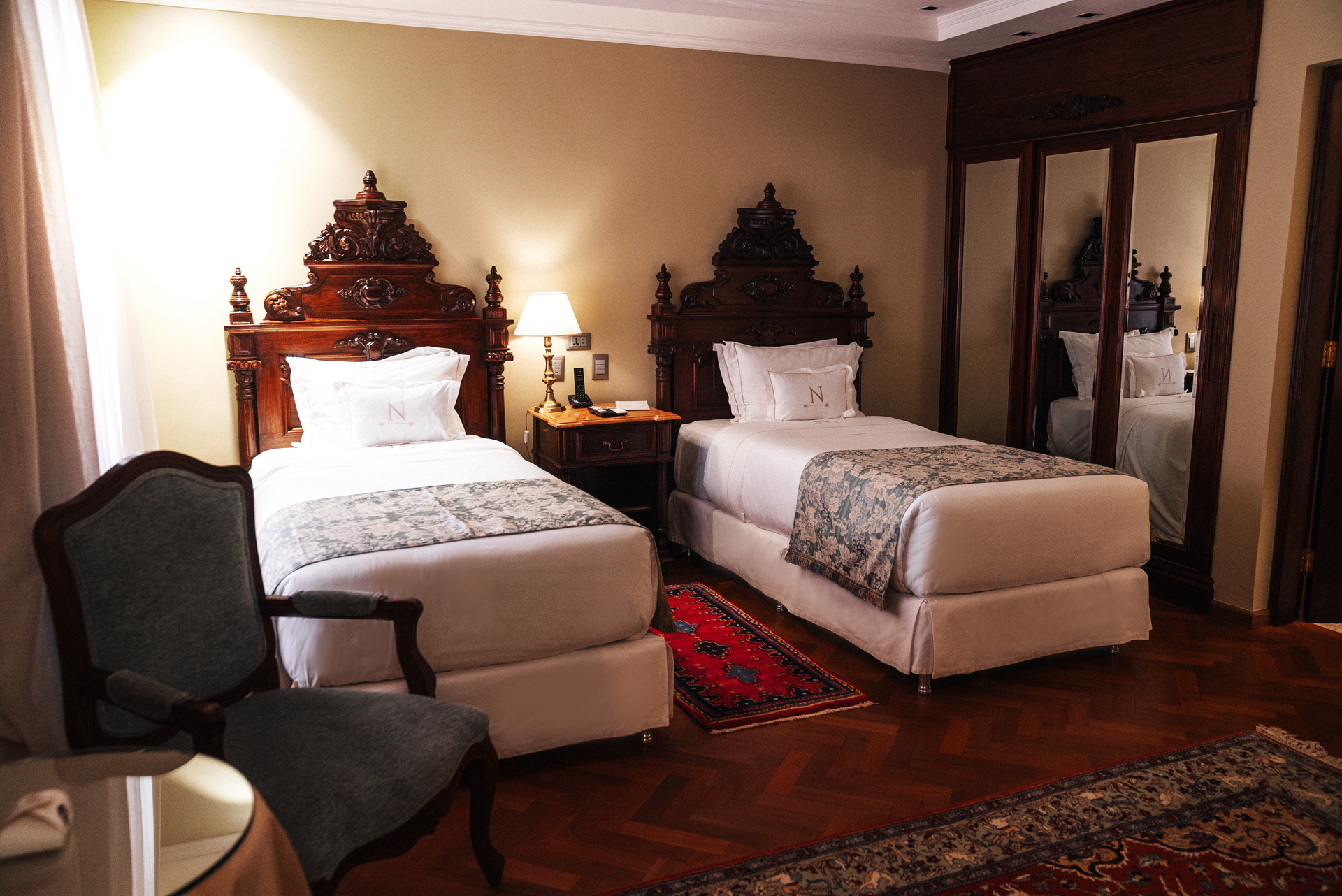Una habitación de hotel con dos camas individuales, cabeceros de madera oscura, una mesita de noche con lámpara, una silla de madera y alfombras estampadas.