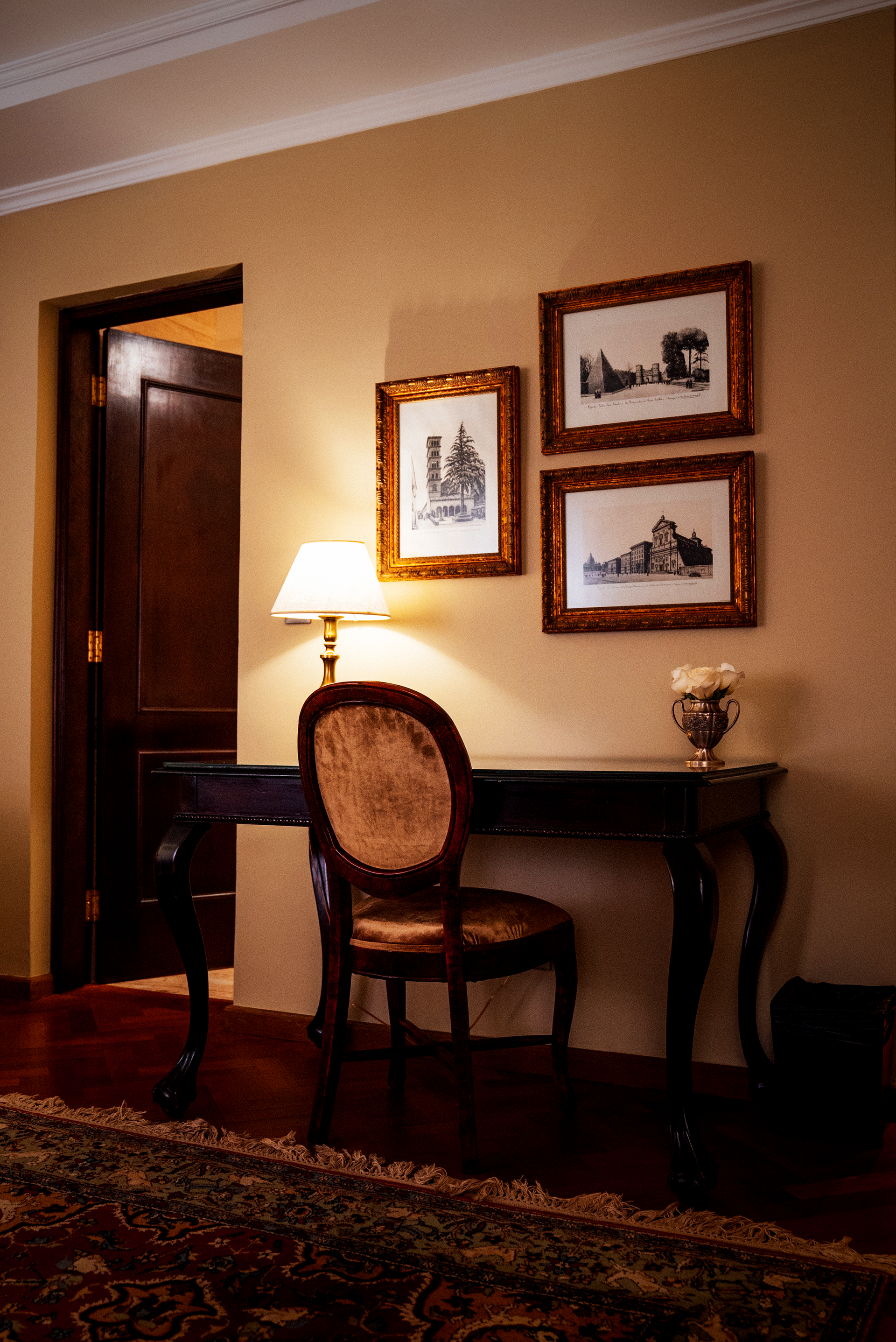 Un escritorio con lámpara, silla y cuadros enmarcados en una pared de color beige entre dos puertas de madera oscura en una habitación con suelo de madera noble.